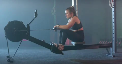 Woman in black bra tights using rowing machine in gym, driving feet pulling Stock Footage 339801695