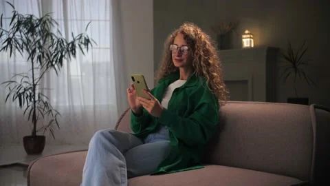 Woman browsing internet using phone sitting at couch.Young curly hair girl Stock Footage 212315614