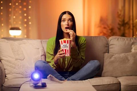 Woman captivated by dramatic film, holding popcorn and staring at screen during Stock Photos