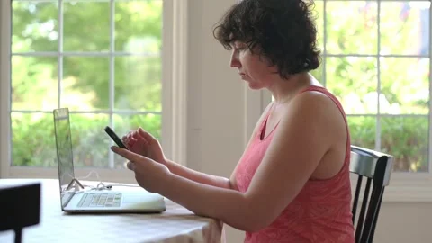 Woman checking phone at kitchen table with laptop, appearing worried or focused Vídeo Stock 320030564
