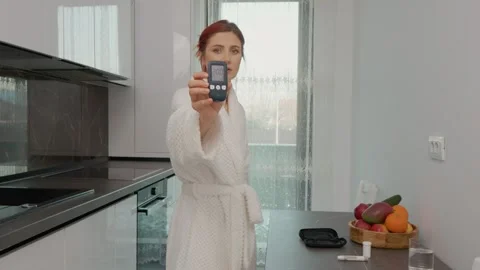 Woman checks glucometer in kitchen with high blood sugar reading Stock Footage 346704200