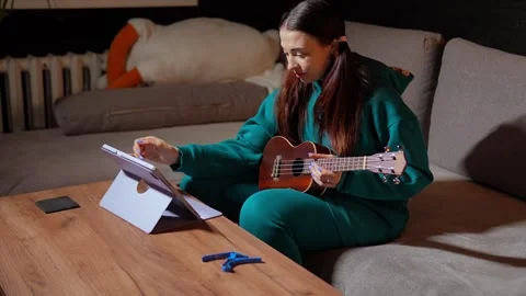 Woman is choosing tutorial online on tablet how to play ukulele sitting with 動画素材 314290551
