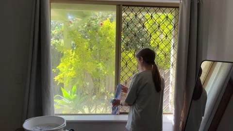 Woman cleans window while sunlight streams through the glass in a home Stock Footage 335063534