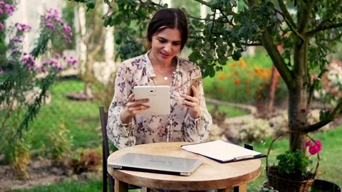 Woman comparing data on smartphone and tablet sitting in beautiful garden Stock Footage 128604360