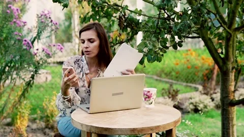 Woman comparing data on smartphone, laptop and tablet sitting in beautiful garde Stock Footage 128602154