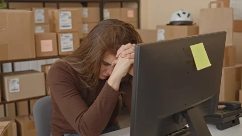 Woman at computer with clasped hands and bowed head among stacked packages .. Stock Footage 337358388