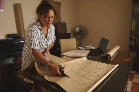 Woman cutting sewing pattern on table in home studio with laptop and sewing.. Stock Photos