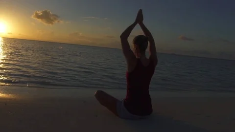 A woman does a seated yoga pose on the beach with hands up at sunset, slow motio Stock Footage