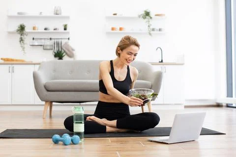 Woman doing yoga exercise while eating healthy salad at home Stock Photos