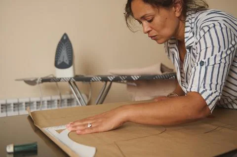 Woman drafting sewing pattern on paper at home workshop with ironing board .. Stock Photos