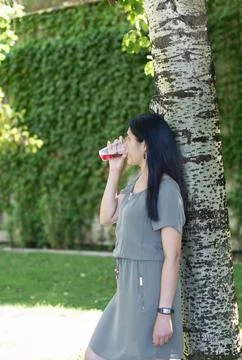 A woman is drinking a beverage while standing next to a tree Stock-Fotos