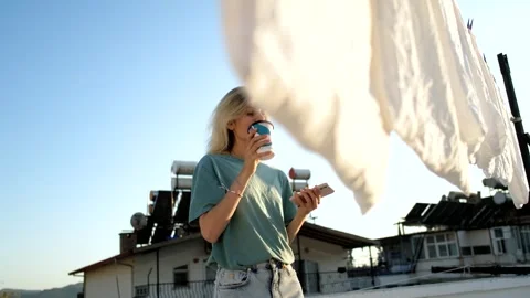 Woman drinking coffee while sitting on the roof of a house with hanging laundry 스톡 동영상 266140173