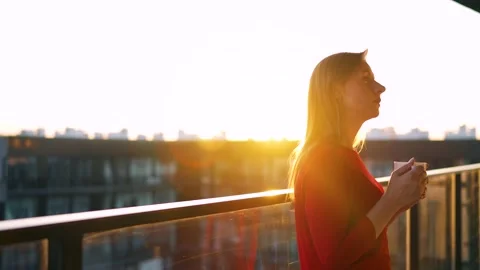 Woman drinking coffee while standing on the balcony and admire the sunset. Slow Stock Footage 197319799