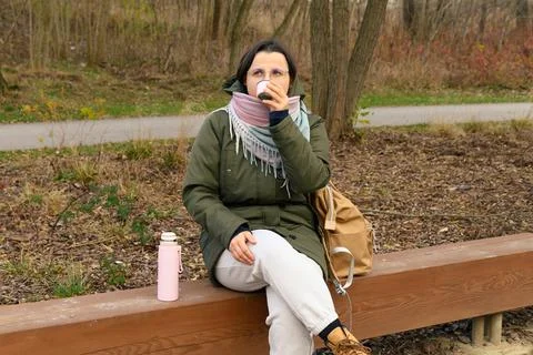Woman drinking from cup while sitting on park bench outdoors Stock Photos