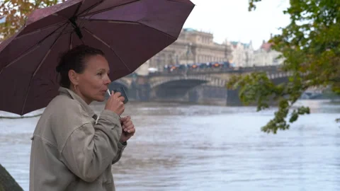 Woman drinking hot beverage under umbrella near river in prague Stock Footage 345113409