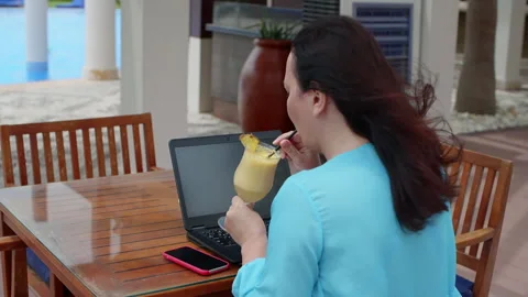 Woman drinking juice while working on laptop at resort on windy day 動画素材 251671148