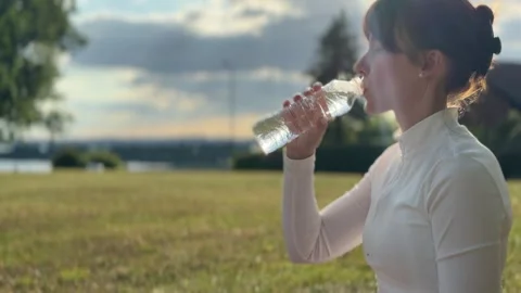 Woman drinking water while sitting on grass in a natural setting. 스톡 동영상 341877588