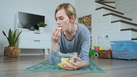 Woman eating chips while standing in the plank at home Stock Footage 143768034