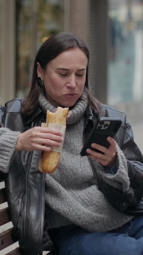 Woman Eating a Sandwich While Using Her Phone on a City Bench Stock-Footage 332402250