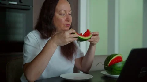 Woman eating watermelon slice while working at laptop in kitchen Stock Footage 339632020