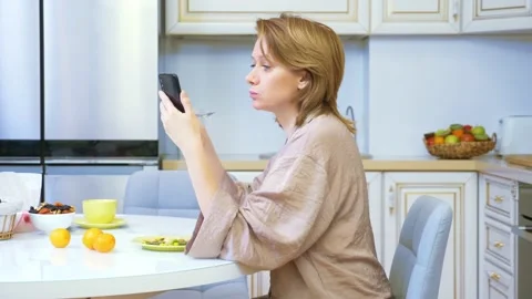 Woman eating while sitting in the kitchen at the table and using her smartphone Stock Footage 145302884