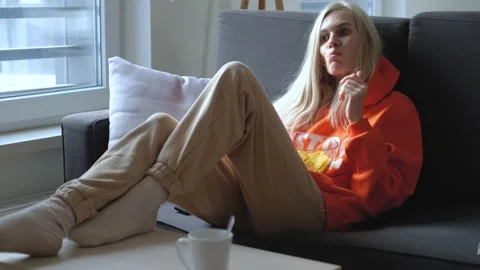 Woman eats chips and turn on tv remote control while sitting on the couch Stock Footage 166518721