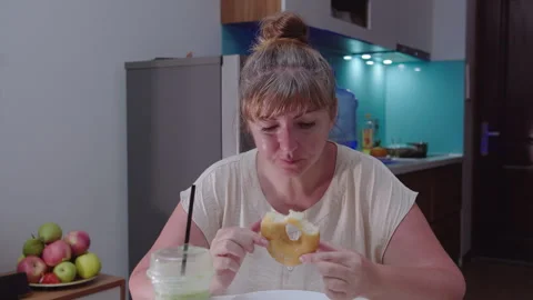 Woman eats a curd ring, sitting at a table, against the background of the Stock Footage 329939164