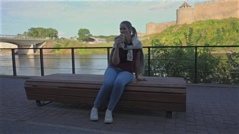 Woman eats ice cream sits on bench by river near castle at sunset Stock Footage 342194547