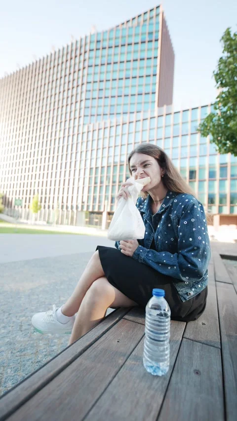 Woman eats a sandwich while sitting on a bench outside a glass office building Stock Footage 348233577