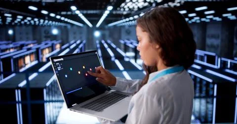 Woman Engineer Monitors Data Servers Using Laptop In Busy Cloud Center. Stock Photos