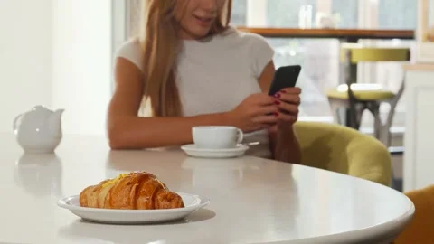 Woman enjoying a croissant while using her smartphone at a cozy bakery café Stock Footage 311545648
