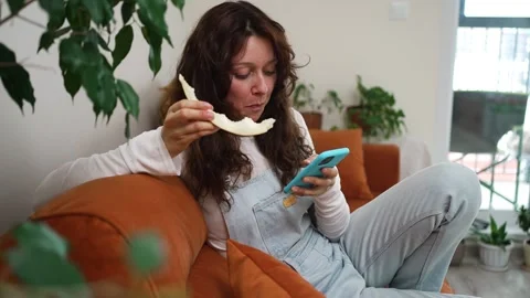 Woman enjoying melon slice while reclining on couch, scrolling smartphone near Stock Footage 315660715
