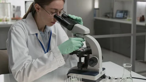 Woman Examining Microscope in Laboratory, with Test Tubes and Beakers on Table Stock Footage 321896959
