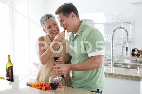 Woman Feeding Pepper To Man In Kitchen Stock Photos