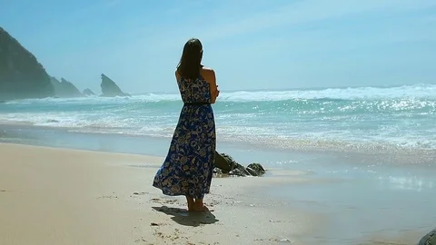 Woman in flowy dress standing on the sandy beach next to the seaside, steadycam Stock Footage