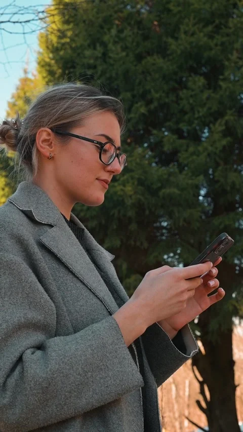 A woman is focused on her smartphone while standing outdoors in a park. The Stock-Footage 337643011