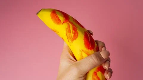 Woman hand holding banana with red lipstick markings on pink background Stock Photos