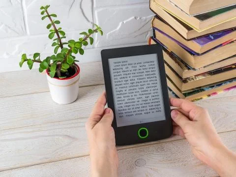 Woman hands hold e-reader near stack of paper books and small potted plant. Stock Photos