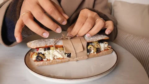 Woman hands untie string on rustic baguette sandwich with cheese and olives. Stock Footage 332897698