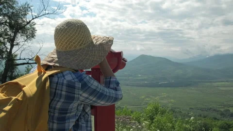 Woman in hat with backpack looking through coin operated binoculars on mountains Stock Footage 230391957