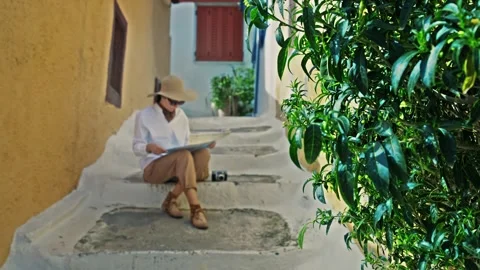 Woman with hat looking at map sitting on staircase of Anafiotika, a part of o 库存影片 276188936