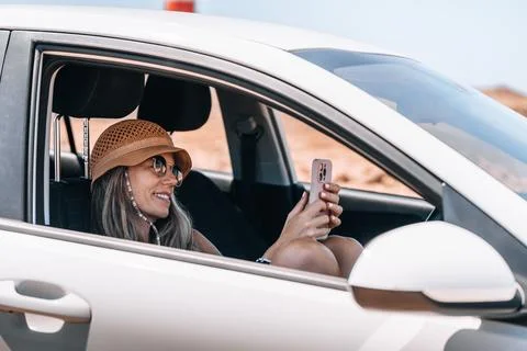 Woman with hat smiles while using her smartphone in a parked car Stock Photos