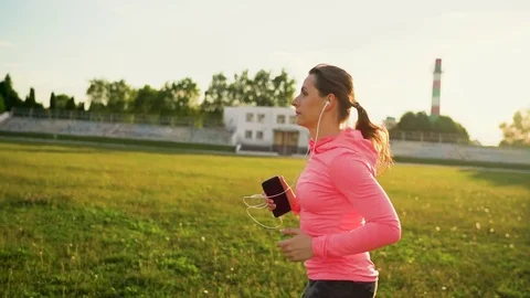 Woman with headphones and smartphone runs through the stadium at sunset Stock Footage