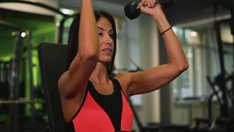 A woman in her 30s performs shoulder presses with dumbbells at a fitness center. 库存影片 345026993
