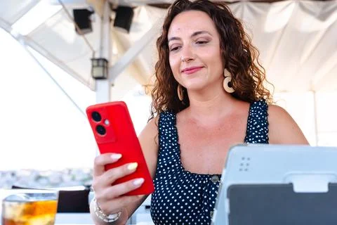 Woman holding red smartphone while using tablet at outdoor cafe workplace Stock Photos