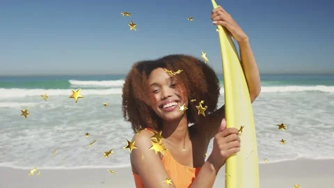 Woman holding yellow board while posing on sandy beach, gold stars appearing and 库存影片 334242536