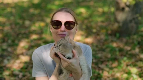 Woman holds cat gently while sitting under trees shade. Vídeo Stock 337228836