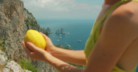 A woman holds a large lemon while standing on a cliff above the Faraglioni sea Stock Footage 345687770