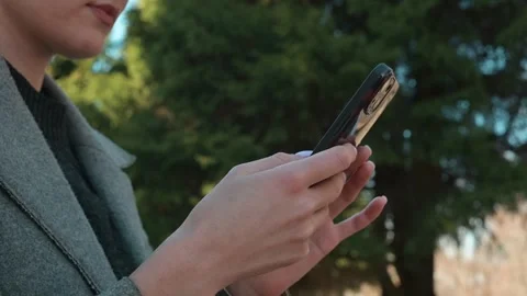 A woman interacts with her smartphone while standing outside in a park. She is 스톡 동영상 337642899