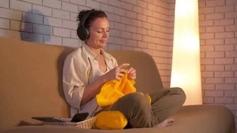 Woman knitting on sofa while listening to music on headphones Video stock 345054348